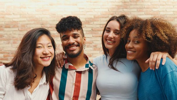 Happy, lovely multiethnic young people posing for the camera on summer day outdoors. Group of friends hugging tightly smiling at camera while standing on brick wall background - Фото, изображение