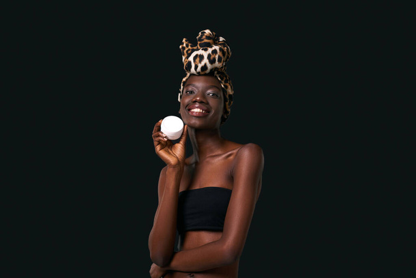 Smiling black girl holding cosmetic cream and looking at camera. Young slim woman wearing traditional african turban and tank top. Body and skin care. Dark background. Studio shoot. Copy space - Fotografie, Obrázek