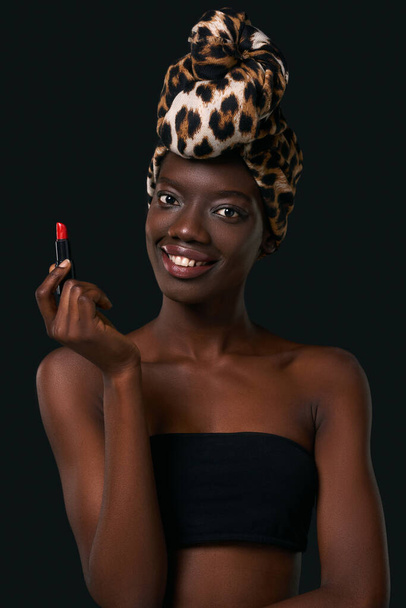 Smiling fashionable black girl holding lipstick and looking at camera. Young slim woman wearing traditional african turban and tank top. Female beauty. Isolated on black background. Studio shoot - Valokuva, kuva