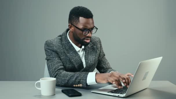 Афроамериканский бизнесмен, работающий в офисе за ноутбуком с кружкой кофе за столом на сером фоне и уставший. Высококачественные 4k кадры - Кадры, видео