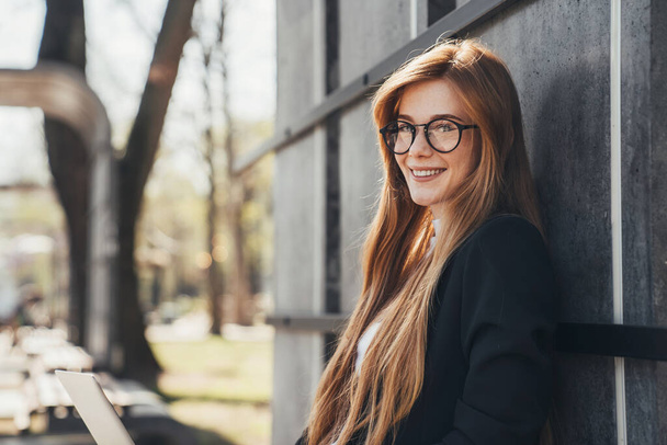 Attractive stylish smiling woman in glasses looking at camera while working typing on laptop, sitting in the park. Smiling Caucasian student studying, learning - Fotó, kép