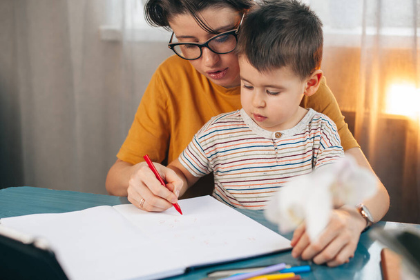 Matka učí svého malého syna psát, zatímco sedí na stole. Obchod, mateřství, multitasking a rodinný koncept. Cvičení psaní, školení - Fotografie, Obrázek