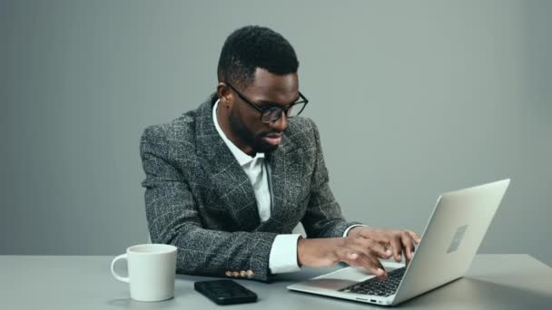 Afro-Amerikaanse zakenman werkt in het kantoor op een laptop met een mok koffie aan zijn bureau op een grijze achtergrond en moe. Hoge kwaliteit 4k beeldmateriaal - Video