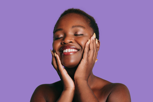Šťastná afroamerická modelka dotýkající se péče o pleť na fialovém pozadí. Etnické přírodní krásy péče o pleť. Koncepce kosmetiky a dermatologie. - Fotografie, Obrázek