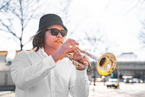 Young European or American male musician jazzman with long hair in white shirt, hat and sunglasses playing on trumpet on sunny town street near road on summer day. Event or concert, masterclass - Foto, afbeelding