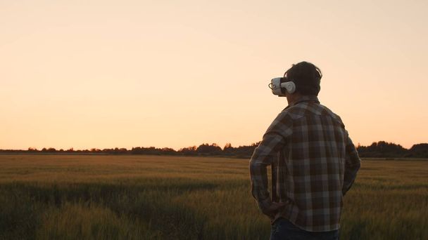 Farmer in virtual reality helmet in front of a sunset agricultural landscape. Man in a countryside field. The concept of country life, food production, farming and technology. - Photo, Image