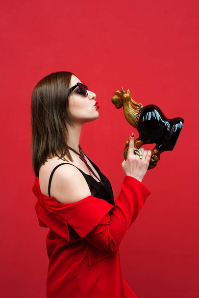 Beautiful girl in a red suit on a red background in profile. In the hands of a figurine cockerel from Bucha, Ukraine - 写真・画像