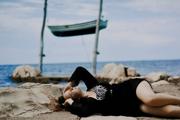 Gorgeous brunette on the beach against the backdrop of the sea and a suspended boat. Woman with long hair resting, summer atmospheric photo. - Valokuva, kuva
