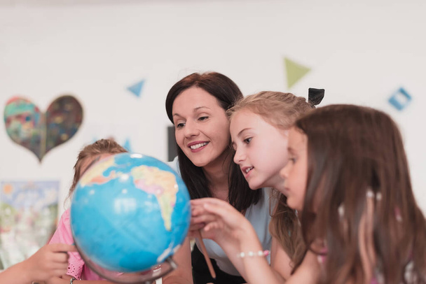 Female teacher with kids in geography class looking at a globe. Side view of a group of diverse happy school kids with a globe in the classroom at school. High quality photo - Foto, Imagen