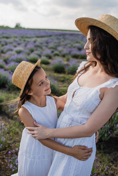 mom and daughter in straw hats smiling at each other in flowering field - Photo, Image