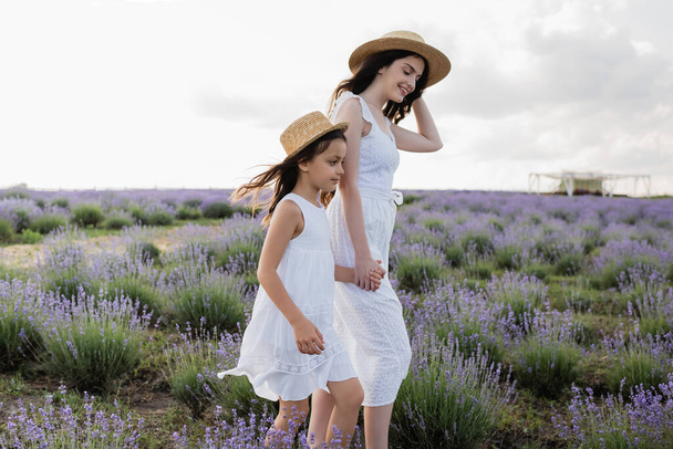 woman and kid in straw hats holding hands while walking in lavender meadow - Photo, image