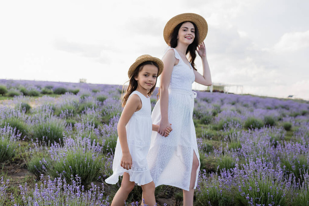 smiling mom and daughter in straw hats holding hands and smiling at camera in lavender field - Foto, Imagen