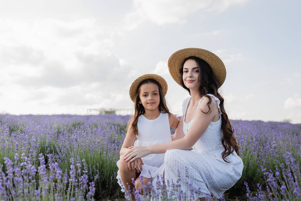 μαμά και παιδί με ψάθινα καπέλα και λευκά φορέματα κοιτάζοντας κάμερα κοντά λεβάντα στον τομέα - Φωτογραφία, εικόνα