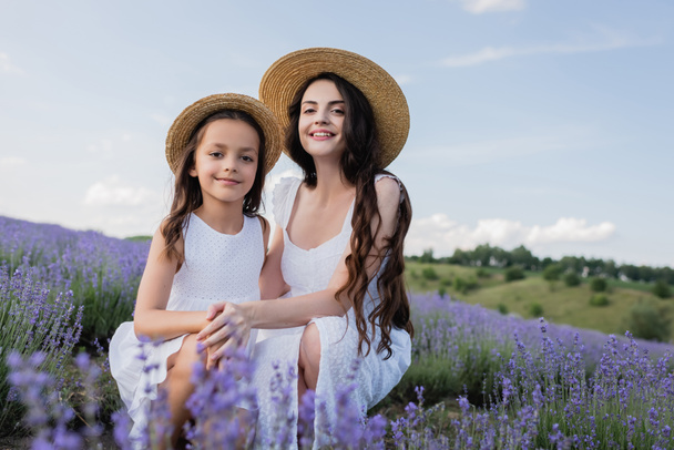 cheerful mom and daughter sitting in lavender field and looking at camera - Foto, Imagen