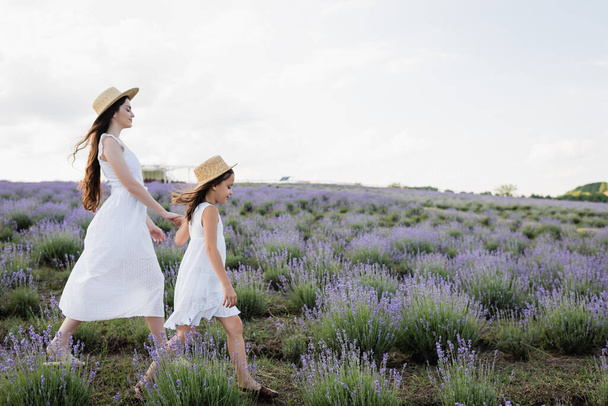 side view of mom and child in white dresses and straw hats holding hands and walking in field - Foto, Imagen
