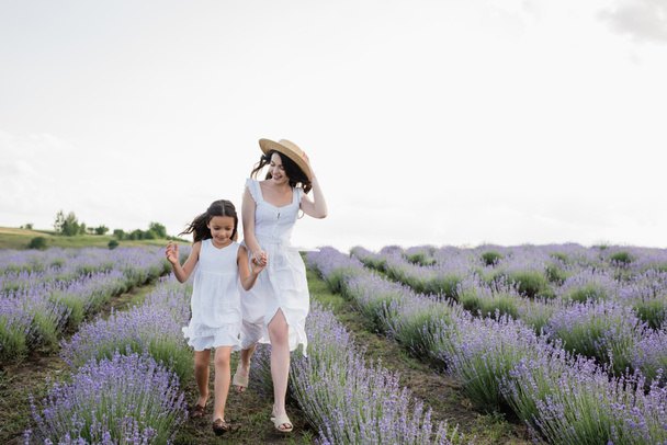 cheerful mother and daughter in white dresses holding hands while running in meadow - Foto, Imagem