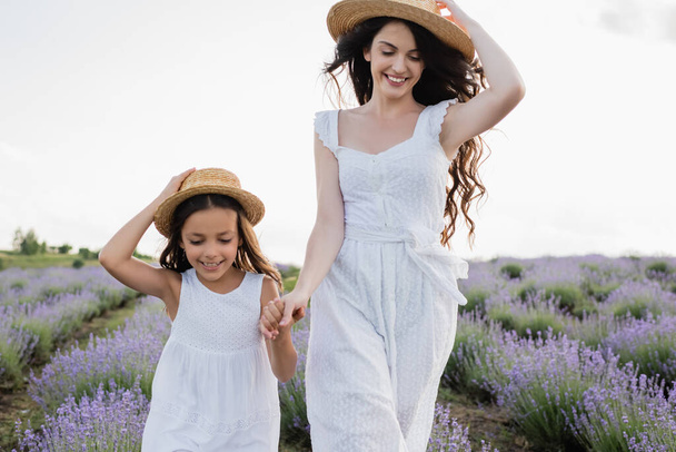 smiling girl and woman in white dresses and straw hats holding hands in field - Fotoğraf, Görsel