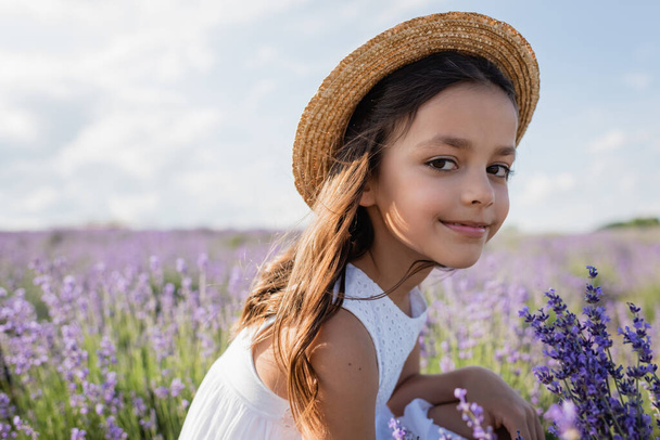 pleased girl in straw hat looking at camera in field with flowering lavender - Foto, imagen