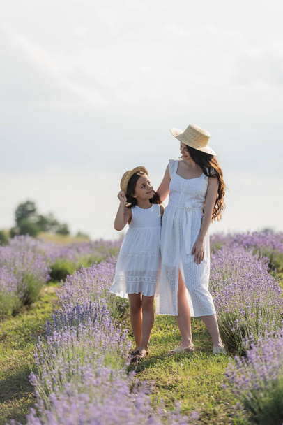 girl and mom in summer dresses and straw hats smiling at each other in blooming meadow - Fotó, kép