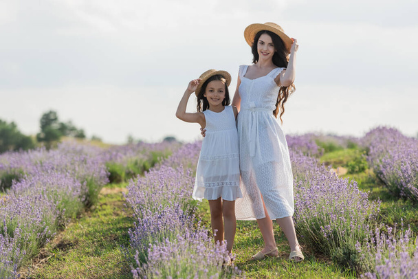 cheerful mom and daughter in straw hats looking at camera in lavender meadow - Foto, Imagen
