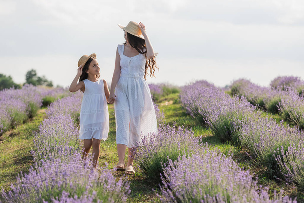 happy mom and daughter in dresses and straw hats holding hands and walking in meadow - Foto, Bild