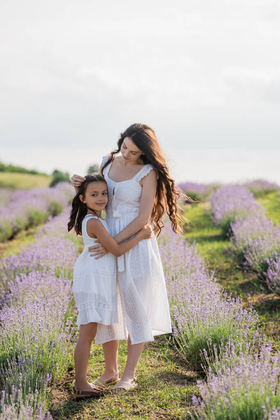 full length of woman and daughter in white dresses embracing in blossoming field - Foto, Imagen