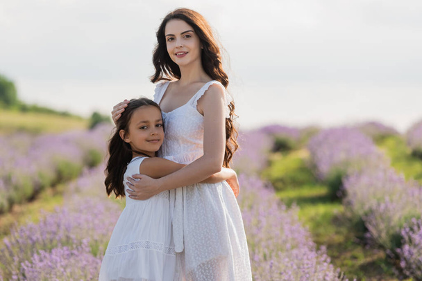 κορίτσι και μαμά σε λευκά φορέματα αγκαλιάζει και κοιτάζοντας κάμερα σε θολή τομέα - Φωτογραφία, εικόνα