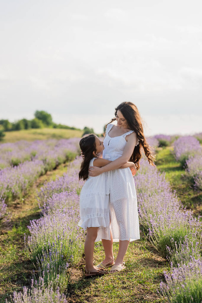 full length of brunette mom and daughter in white dresses embracing in meadow - Foto, Imagen