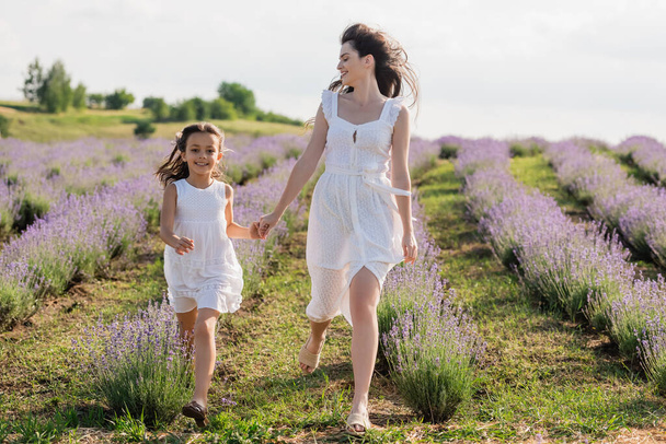 cheerful woman and girl in white dresses holding hands and running in field - Fotó, kép