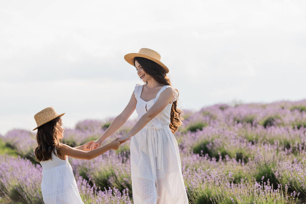 joyful mom and daughter in straw hats holding hands while having fun in field - Photo, Image