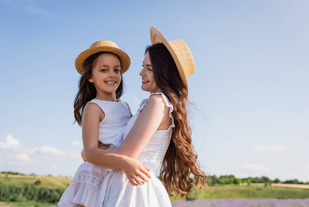 cheerful girl in straw hat looking at camera in hands of mom outdoors - Foto, Imagen