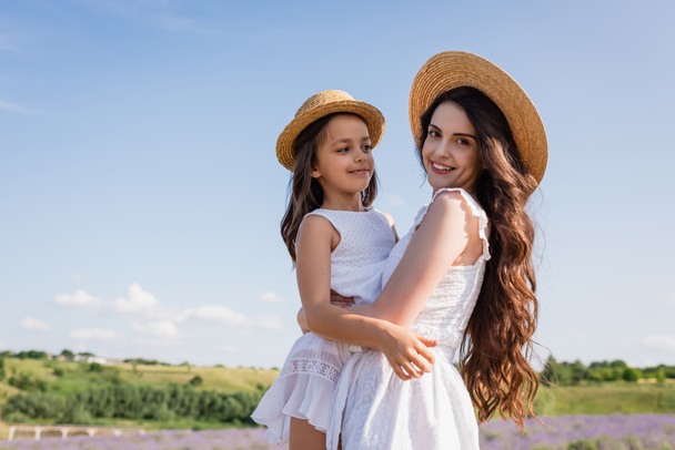 brunette woman in straw hat smiling at camera while holding daughter in countryside - Photo, Image