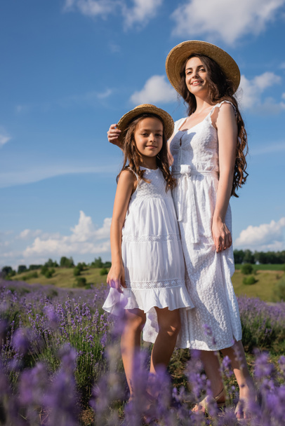 Низкий угол зрения мамы и дочери в белых платьях, стоящих в цветущем поле - Фото, изображение