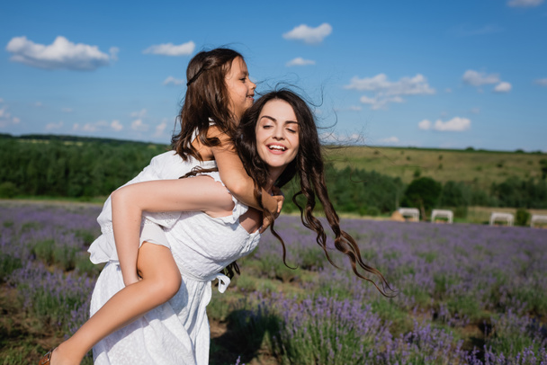 cheerful brunette woman piggybacking daughter in flowering field - Foto, afbeelding