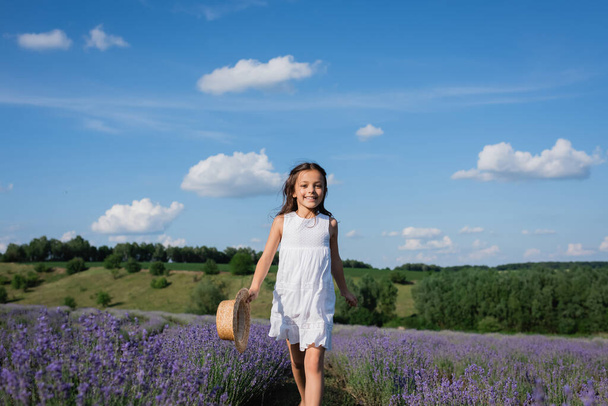 χαρούμενο κορίτσι με καλοκαιρινό φόρεμα περπατώντας στον τομέα της λεβάντας κάτω από τον γαλάζιο ουρανό με λευκά σύννεφα - Φωτογραφία, εικόνα