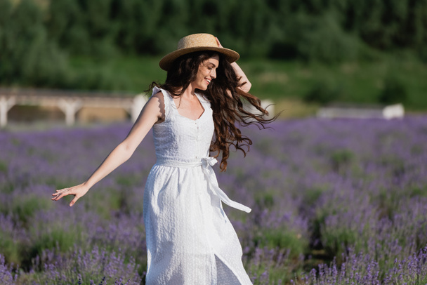 happy brunette woman in white dress and straw hat walking in field with blossoming lavender - Foto, Imagem