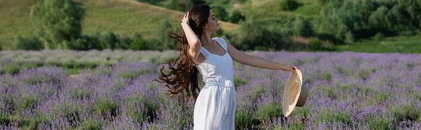 woman in white dress touching long hair and holding straw hat in lavender field, banner - 写真・画像