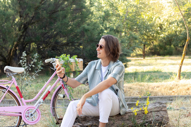 Молодий чоловік сидить на дерев'яному багажнику, надсилаючи повідомлення своїм друзям на смартфон з рожевим вінтажним велосипедом і рослинами на задньому плані
 - Фото, зображення