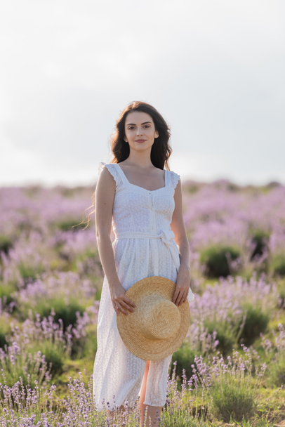 smiling woman in white dress holding straw hat and looking at camera in lavender meadow - Foto, imagen