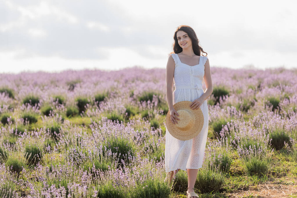 happy woman in summer dress holding straw hat while standing in lavender field - Foto, afbeelding