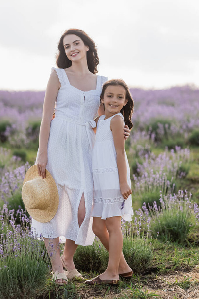 happy mother and daughter in white summer dresses smiling at camera in field - Photo, image