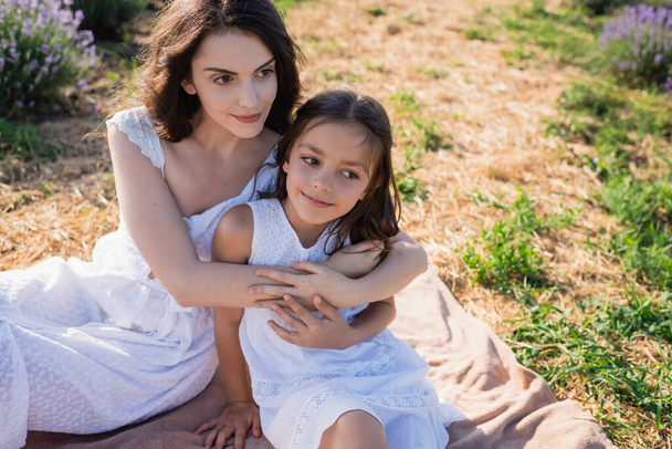 happy brunette woman embracing daughter while sitting in field on summer day - Photo, image