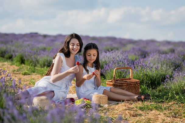 smiling mother and child eating strawberries during picnic in lavender meadow - Photo, image