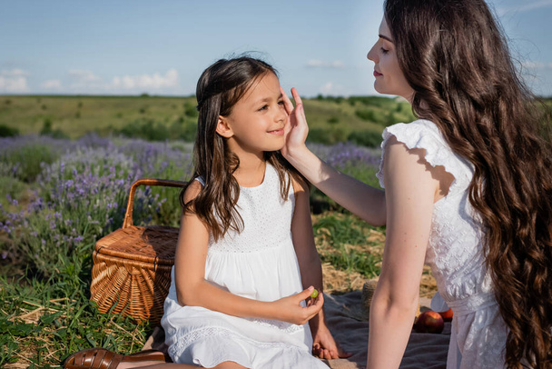 happy woman touching face of daughter while having picnic in meadow - Photo, image