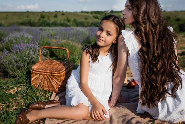 happy girl looking at camera near mom and wicker basket in field - Photo, image