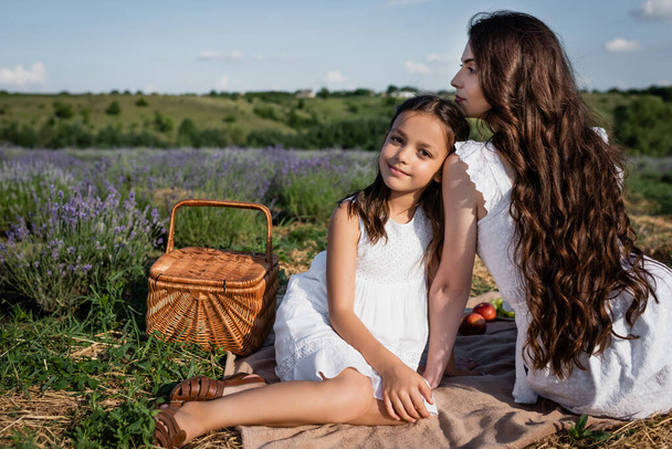 joyful girl looking at camera near brunette mom and straw basket in lavender meadow - Fotó, kép