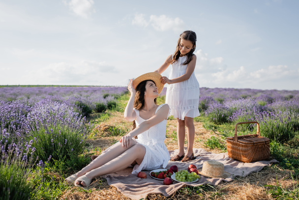 girl holding straw hat near happy mom sitting on blanket near fresh fruits - Fotografie, Obrázek