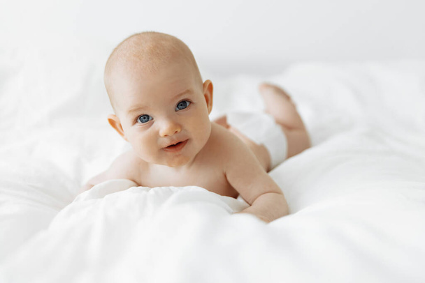 Smiling newborn baby on a white bed at home in a white sunny bedroom. Textiles and bed linen for children. Happy healthy baby concept, space for text - Foto, imagen