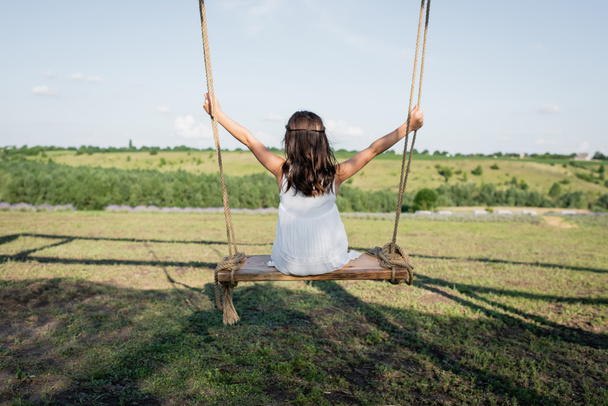 back view of child in summer dress riding swing in meadow - Foto, Imagem