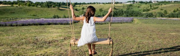 back view of kid in white summer dress riding swing in meadow, banner - 写真・画像